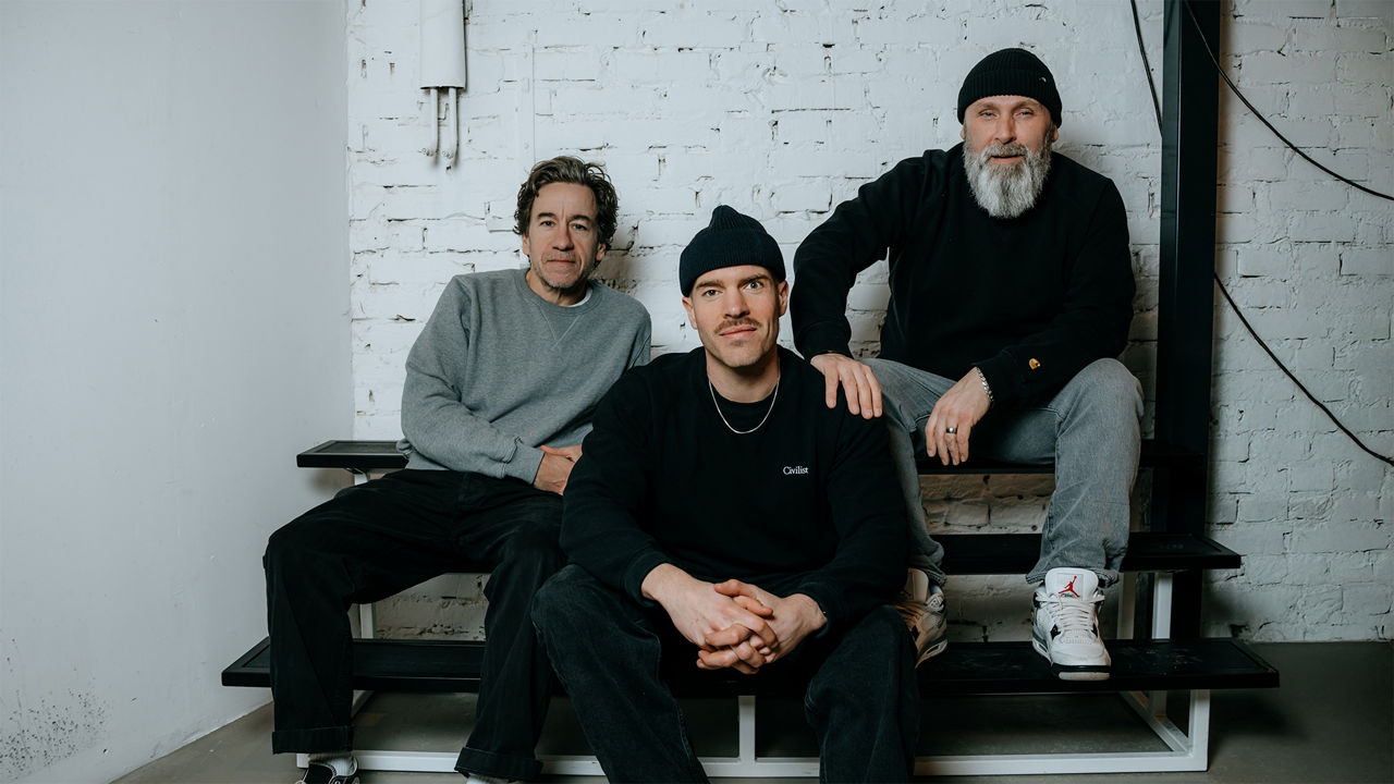 Drei Personen sitzen auf einer schwarzen Sitzbank vor einer weißen Backsteinwand und blicken in die Kamera.