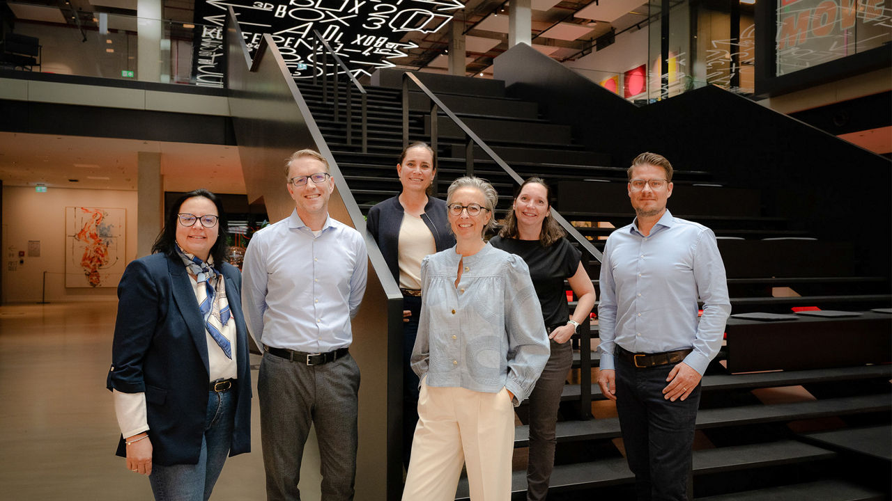 Sechs Personen aus dem Team von Mediaplus und der TARGOBANK stehen im lichtdurchfluteten Atrium des House of Communication in München vor einer schwarzen Tribünen-Treppe. Die Gruppe, bestehend aus drei Frauen und drei Männern, lächelt in die Kamera. Im Hintergrund ist moderne Architektur mit offenen Etagen, Glasfronten und einer auffälligen Lichtinstallation mit typografischen Elementen zu sehen. Die Kleidung ist überwiegend business casual. Die Aufnahme vermittelt ein Gefühl von Zusammenarbeit und Innovation.