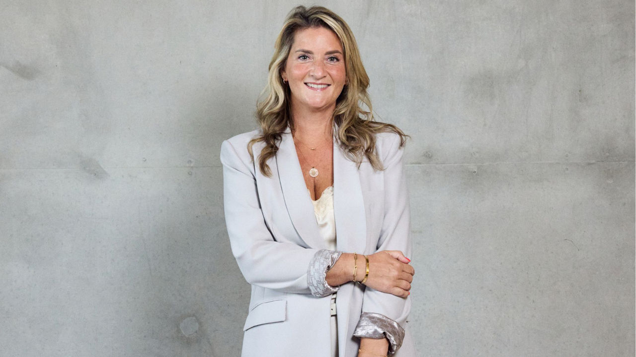 Professional portrait of a person with shoulder-length hair wearing a light-coloured blazer and top, standing against a light grey concrete wall. The person is facing the camera with arms loosely crossed. The image is presented in a business portrait style with a neutral background.