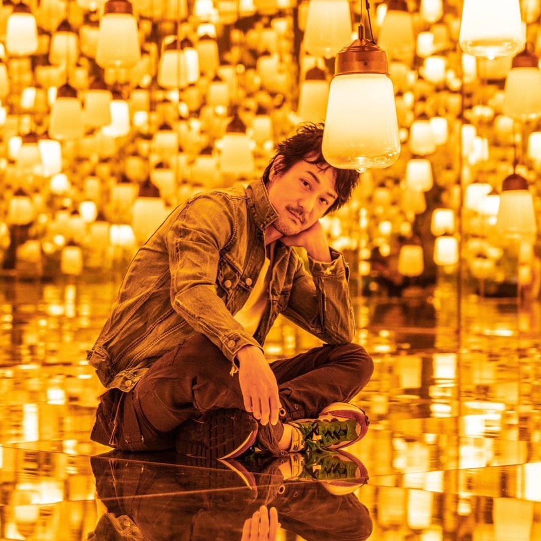 Mann sitzt in einem Raum voller warm leuchtender Lampen, die den Raum spiegelnd erhellen. Die Szene wirkt immersiv und künstlerisch inszeniert.