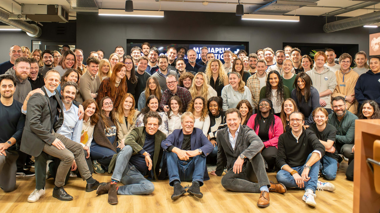 A large and diverse group of people, including men and women of various ages and ethnicities, are gathered in an office space, smiling and posing for a team photo, with some seated on the floor and others standing in front of a dark wall with illuminated signage in the background.