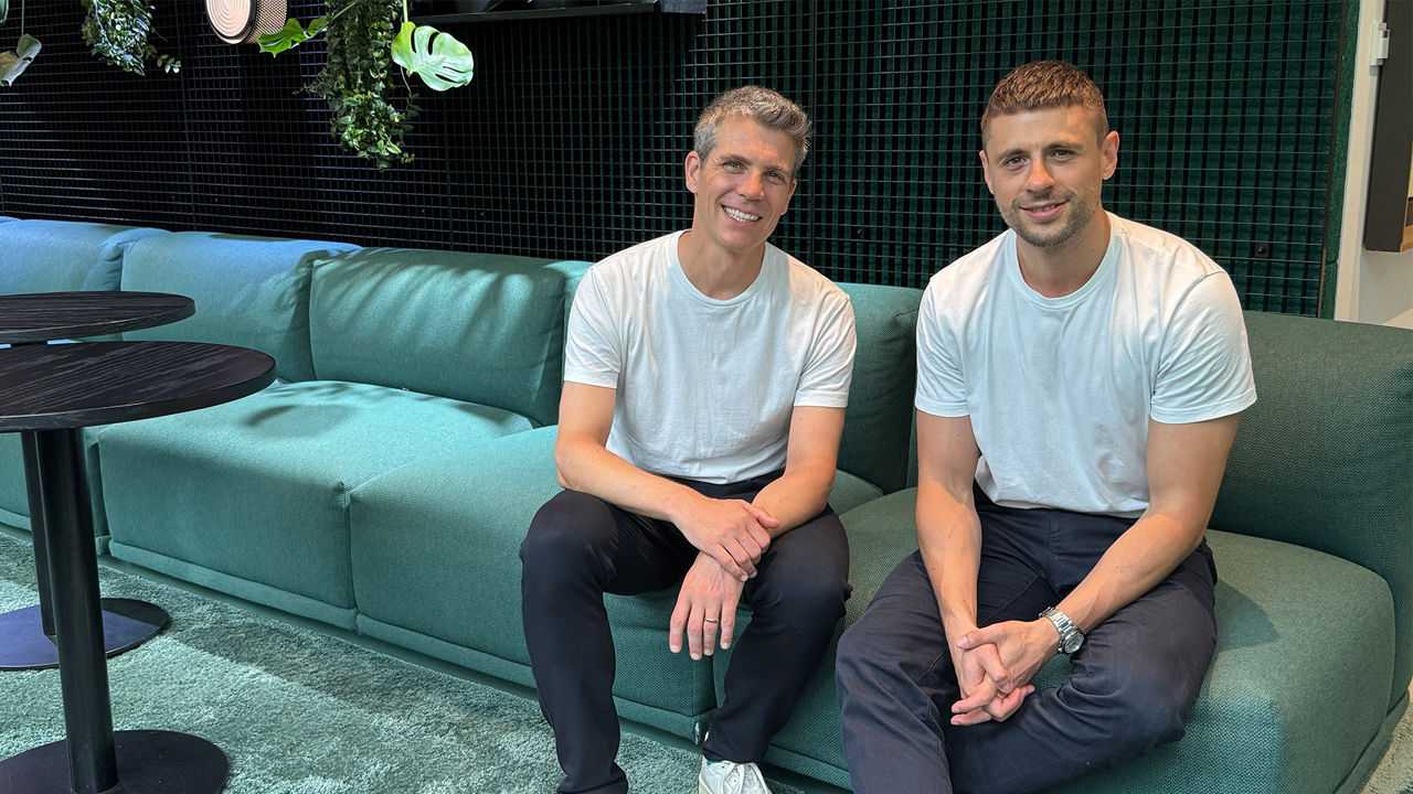 TWELVE CAST mit Benjamin Majeron und Alexander Nagel: Zwei Männer sitzen nebeneinander auf einem modernen, türkisgrünen Sofa in einem Büro mit stilvollem, dunkelgrünem Wandgitter und hängenden Pflanzen. Beide tragen weiße T-Shirts, schwarze Hosen und helle Sneakers. Der Mann links ist etwas älter, hat graues, kurzes Haar und lächelt freundlich in die Kamera. Der Mann rechts hat dunkleres, kurz geschnittenes Haar und trägt eine silberne Armbanduhr. Er sitzt mit verschränkten Händen und entspanntem Gesichtsausdruck. Die Szene wirkt freundlich und offen. Das Foto wurde bei natürlichem Licht aufgenommen.