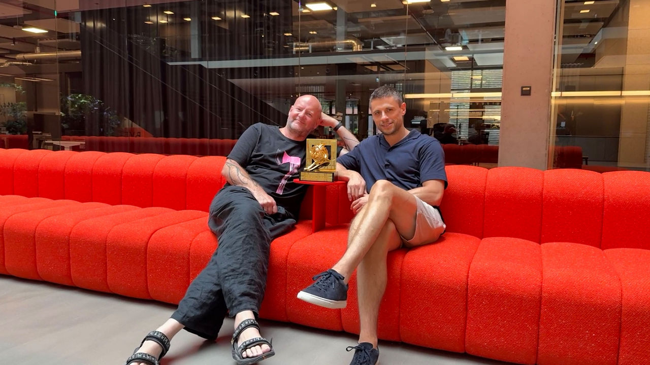 TWELVE CAST mit Benjamin Majeron und Christoph Everke. Zwei Männer sitzen entspannt auf einem leuchtend roten Sofa in einem modernen Bürogebäude. Zwischen ihnen steht ein goldener Preis in Form eines Löwen – ein Cannes Lions Award. Beide Männer lächeln in die Kamera; der linke trägt ein schwarzes Shirt und weite Hose mit Sandalen, der rechte ein dunkelblaues Poloshirt mit kurzen, hellen Shorts und Sneakern.