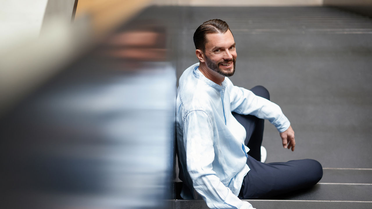 Porträtaufnahme einer Person mit hellblauem Hemd, die auf einer Treppe in einem modernen Gebäude sitzt. Die Person lehnt an einer Wand und blickt in Richtung Kamera. Der Vordergrund ist teilweise unscharf, wodurch die Treppe und die Person hervorgehoben werden. Das Bild ist als professionelles Business-Porträt gestaltet.
