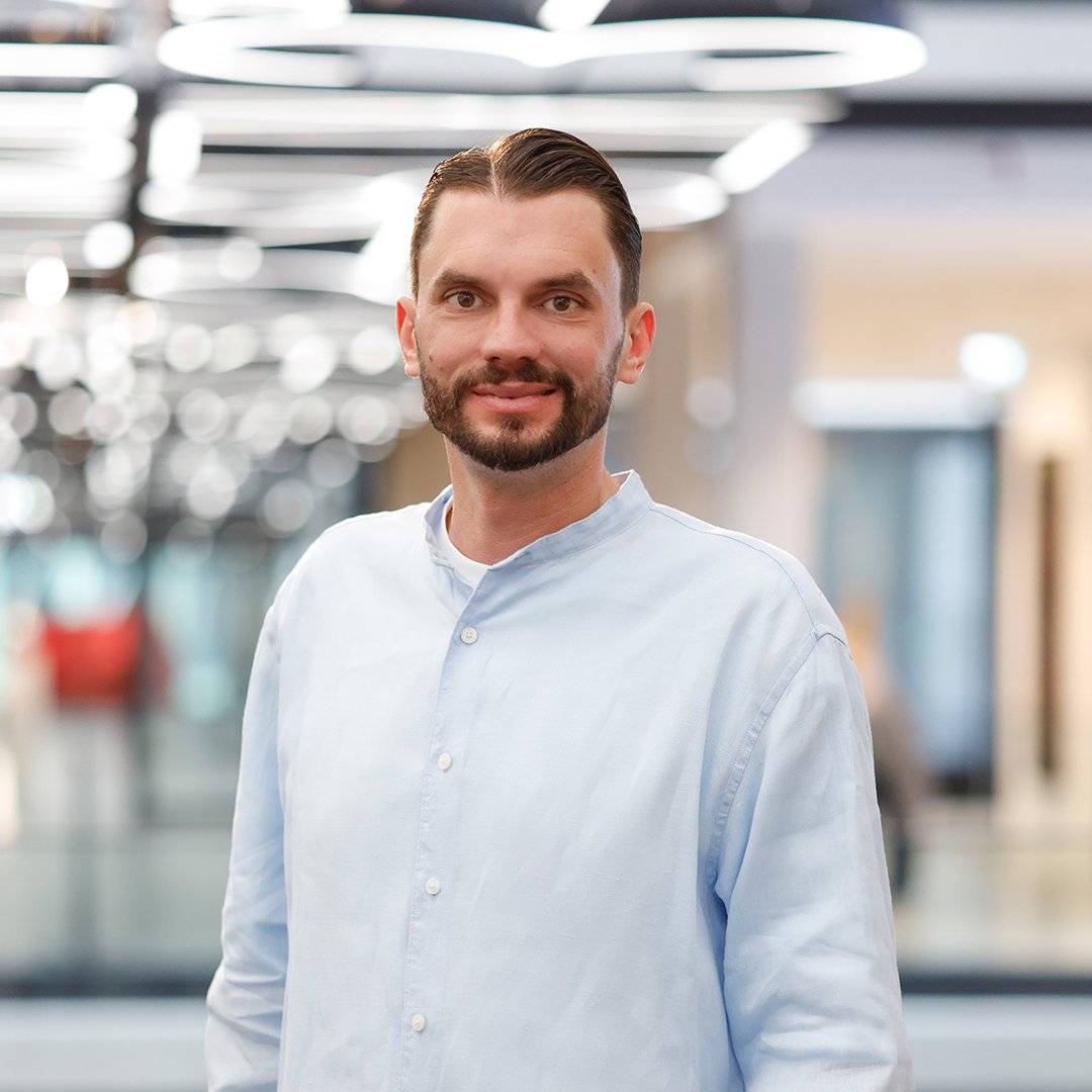Professional portrait of a person wearing a light blue button-up shirt, standing in a modern office setting. The background is softly blurred and features circular ceiling lights and office interiors. The person is facing the camera. The image is presented in a business portrait style.