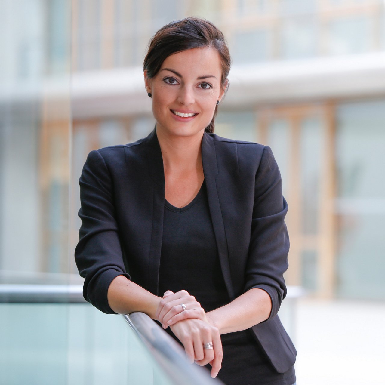 Eine lächelnde Frau in einem schwarzen Blazer lehnt sich an ein Geländer in einem modernen Bürogebäude und blickt direkt in die Kamera.