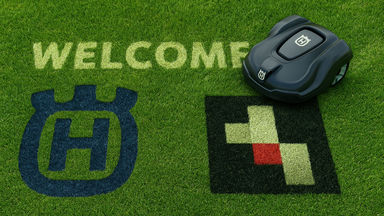 Ein automatischer Rasenmähroboter der Marke Husqvarna steht auf einer Rasenfläche, auf der das Wort „WELCOME“ sowie die Logos von Husqvarna und der Serviceplan Group in den Rasen gemäht sind.