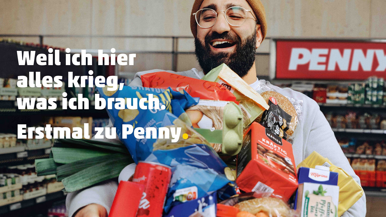 Eine Person mit mehreren Lebensmitteln in den Armen steht in einem Penny-Markt. Im Bild ist ein Werbeslogan von Penny eingeblendet