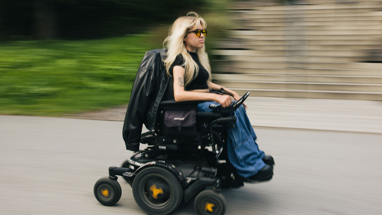 A person with long light-colored hair drives an electric wheelchair along an outdoor path. Motion blur in the background emphasizes movement, while a staircase is visible in the distance.