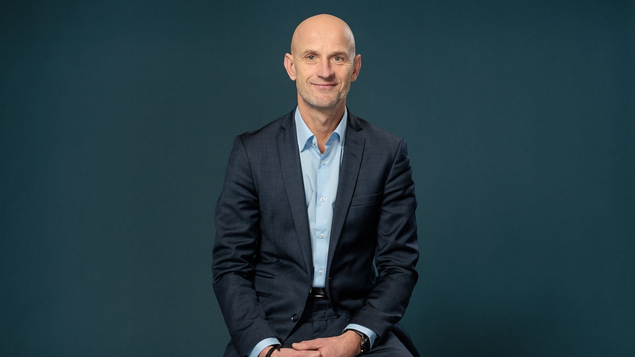 John Wittesaele, COO International Markets bei Mediaplus Group: Das Bild zeigt einen glatzköpfigen Mann mittleren Alters mit hellem Hauttyp, der in einem dunkelblauen Anzug mit hellblauem Hemd vor einem einfarbigen, dunklen Hintergrund sitzt, leicht lächelt und direkt in die Kamera blickt.