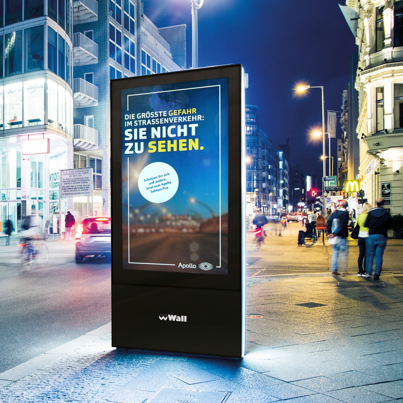 Nighttime street scene in a city with an illuminated Apollo Optik billboard on the side of the road. The sign reads: 'The biggest danger on the road: not being able to see you. Protect yourself and others. Now for the Apollo Eye Test Pro.