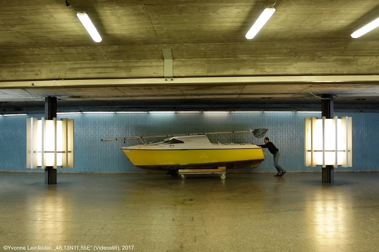 Artwork: "48, 13N 11, 58E" by Yvonne Leinfelder 2017. It shows a small yellow boat being pushed through a warehouse by a man