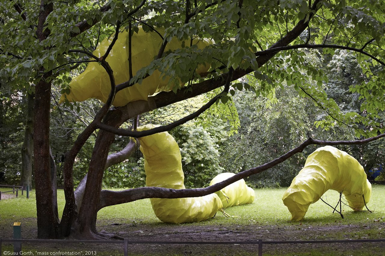 Featured work of art on the talk show "Artpros": mass confrontation by Susu Gorth, 2013. Depiction of oversized, yellow caterpillars on and next to a tree in the park