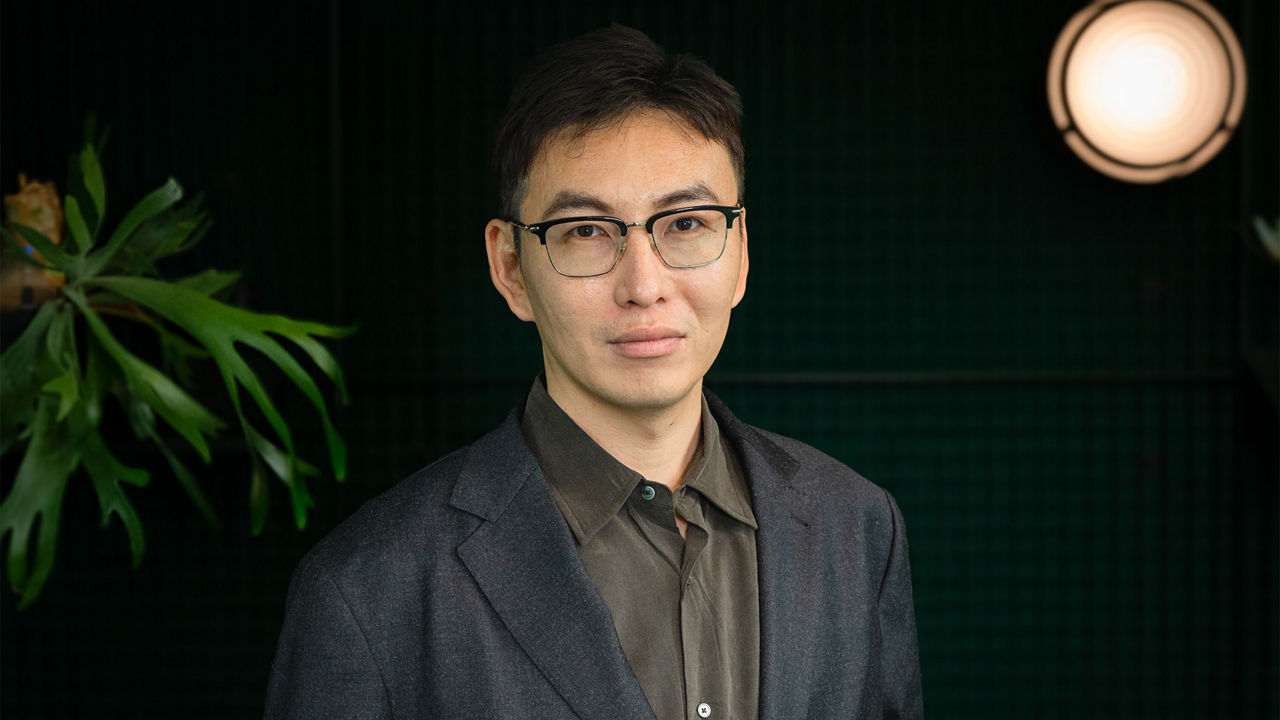Portrait of a person wearing glasses, a dark blazer, and a shirt in front of a dark background. A decorative plant and a round light fixture are visible in the background. The person is facing the camera. The image is presented in a professional business portrait style.