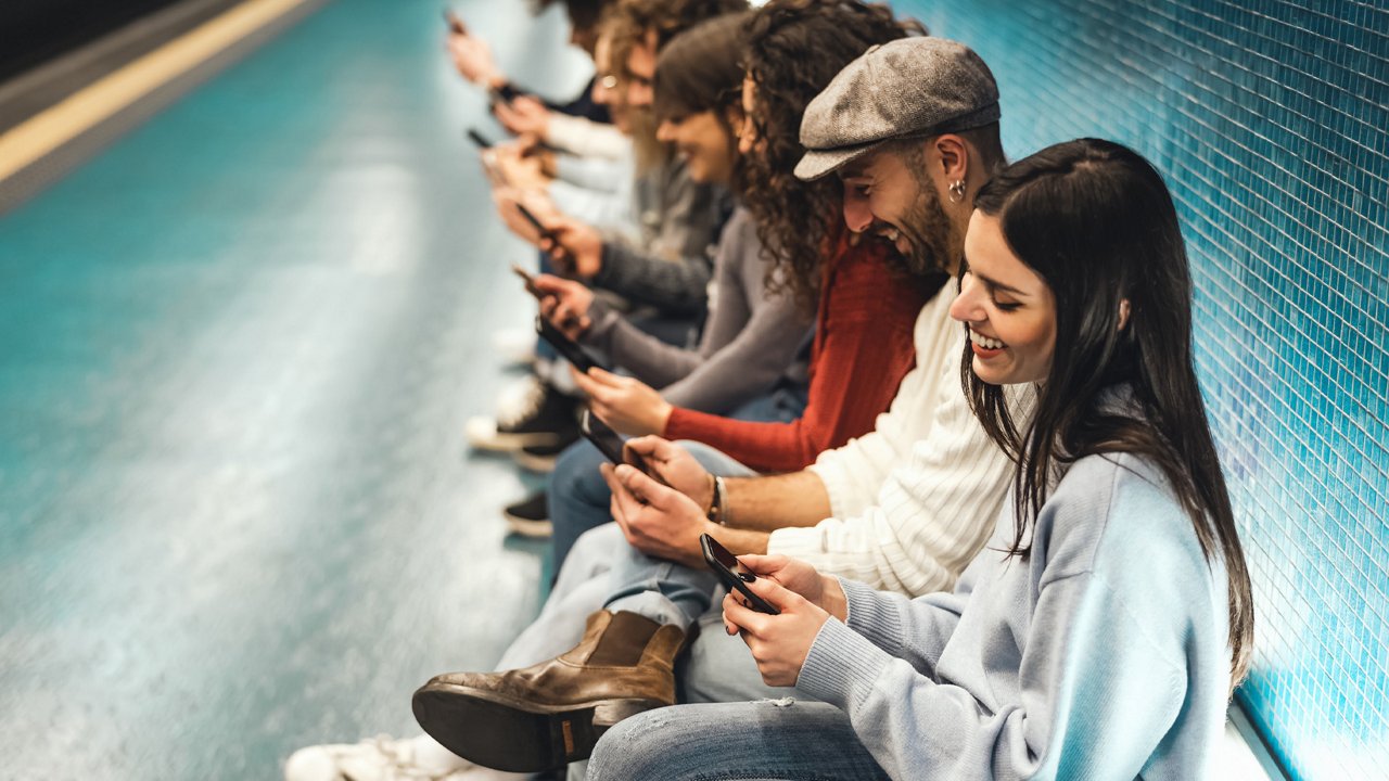 Row of young people sitting against a tiled wall, all looking at their smartphones