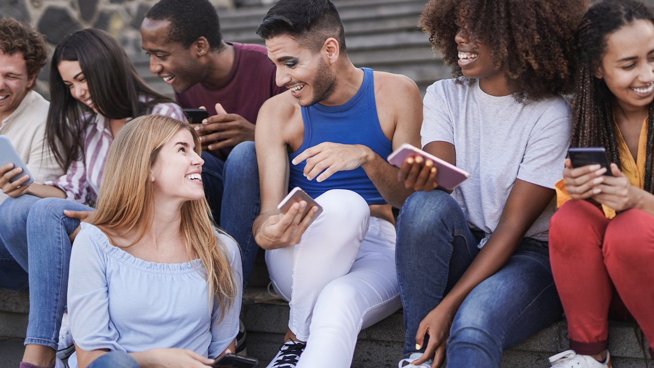 Diverse group of young people sitting on outdoor steps, laughing and sharing content on their phones