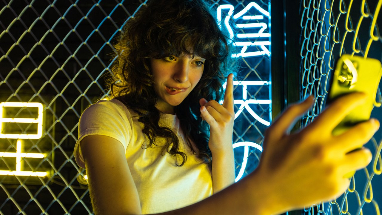 Young woman taking a selfie with a yellow phone in front of a neon-lit chain-link fence