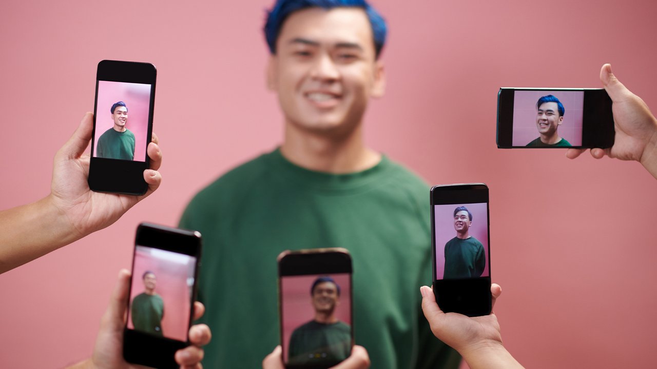 Young man with blue hair being photographed simultaneously by multiple smartphones against a pink background