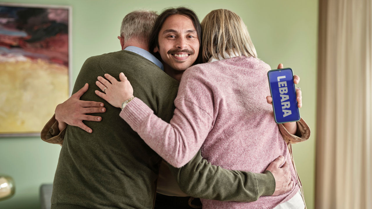 People hugging. One guy facing the the camera is showing a smartphone.