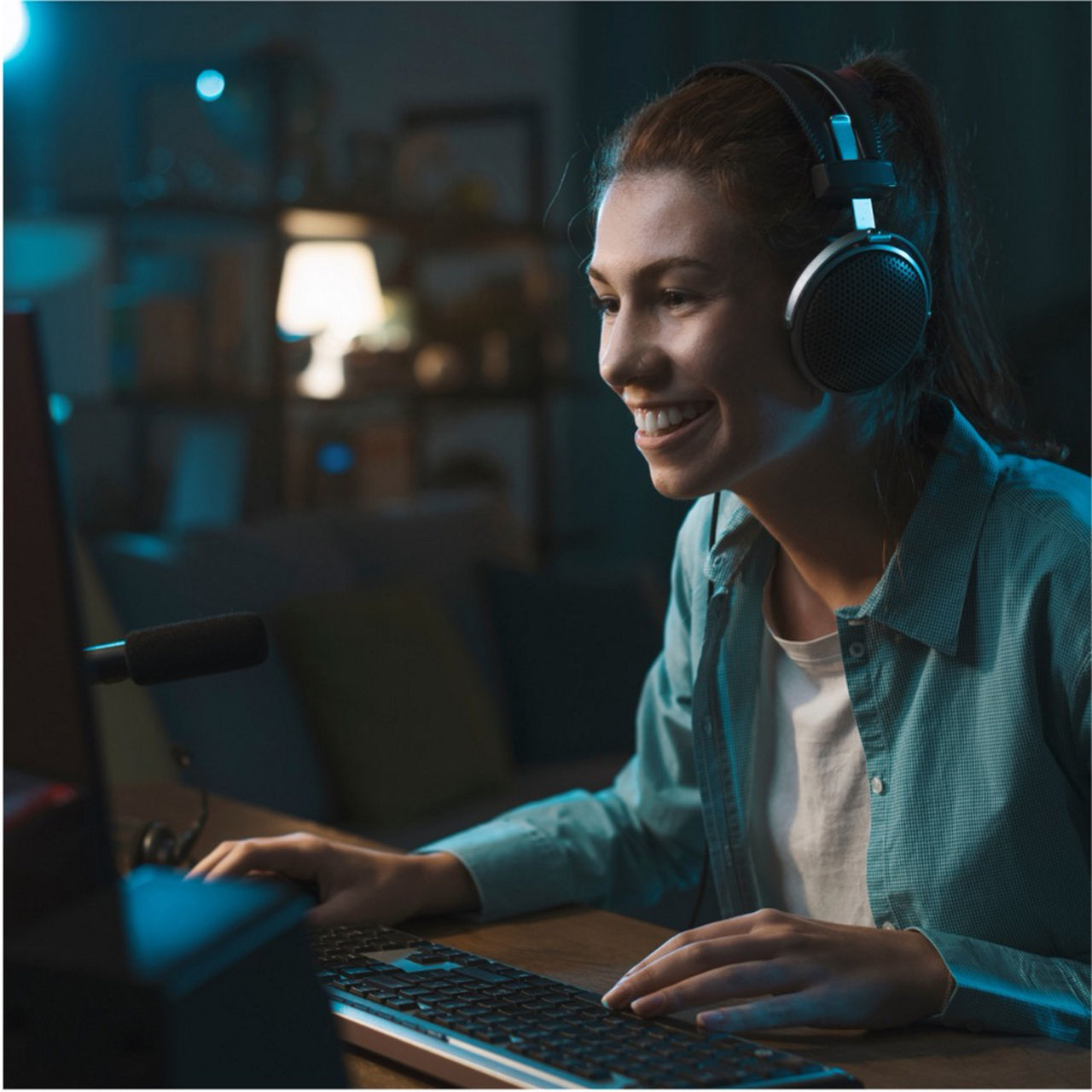 A young woman wearing headphones smiles while using a computer and microphone, suggesting online communication or streaming