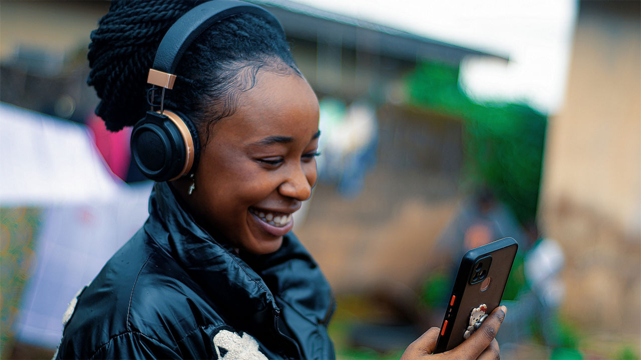 A smiling woman wearing headphones looks at her smartphone outdoors; the background is blurred, suggesting a lively environment.