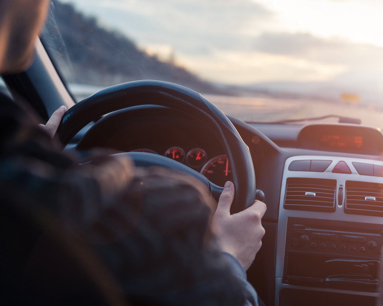 Person driving a car with hands on the steering wheel, view of the dashboard and road ahead.