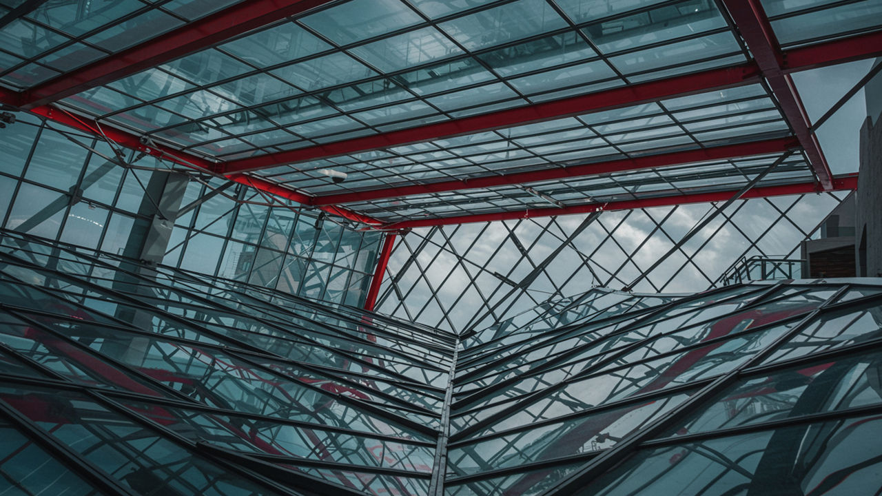 A young woman with short dark hair stands by a large office window, gazing thoughtfully into the distance, a red notebook resting on the windowsill beside her