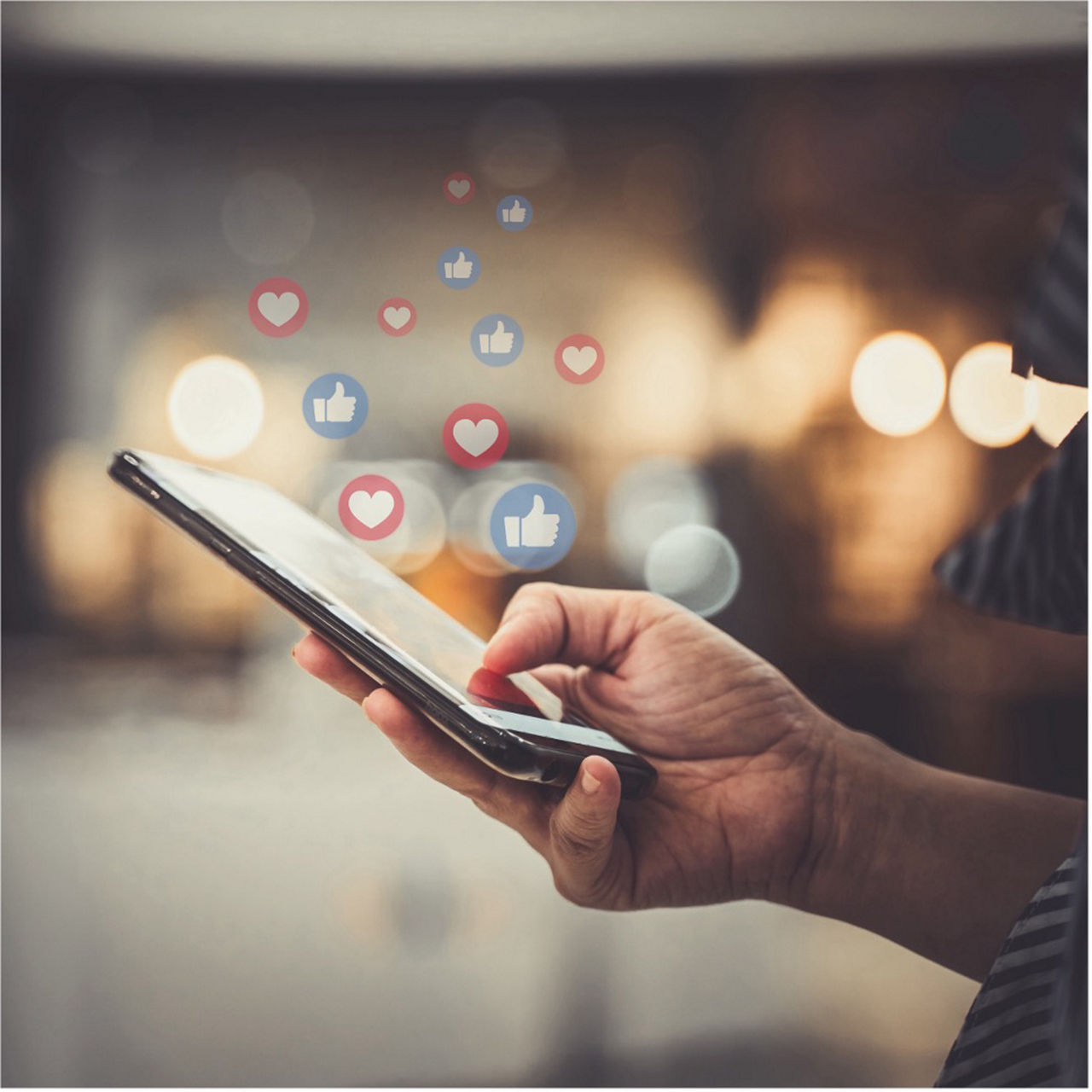A hand holds a smartphone with floating heart and thumbs-up icons, symbolizing social media engagement and interaction