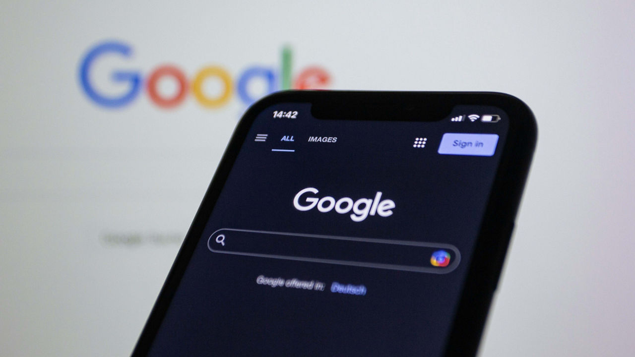 A smartphone screen displays the Google search homepage in dark mode, with the search bar and ‘Sign in’ button clearly visible. In the blurred background, the colorful Google logo appears on a desktop screen.