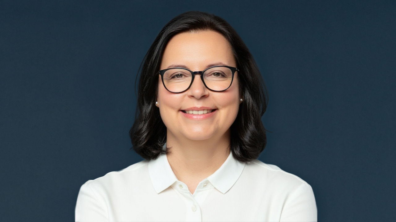 Porträt einer Frau mit dunklem, schulterlangem Haar, schwarzer Brille und weißer Bluse. Sie blickt mit freundlichem Ausdruck in die Kamera. Der Hintergrund ist in dunklen Blautönen gehalten und lenkt den Fokus auf die Person.