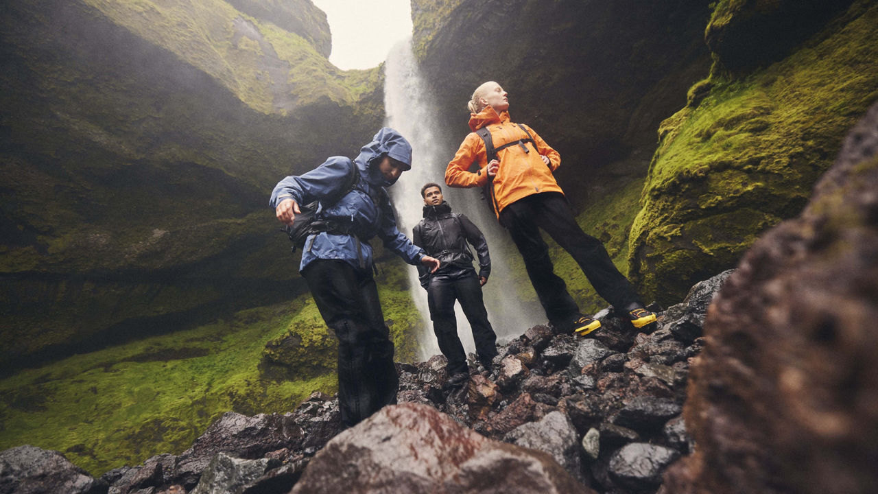 Drei Wandernde in wetterfester Outdoor-Kleidung stehen auf nassen, rutschigen Felsen vor einem hohen Wasserfall in einer moosbedeckten Schlucht. Eine Person trägt eine blaue Jacke mit Kapuze, eine andere eine orange Jacke und steht mit erhobenem Blick. Die dritte Person in dunkler Kleidung steht zwischen ihnen. Die Szene wirkt abenteuerlich und naturverbunden.