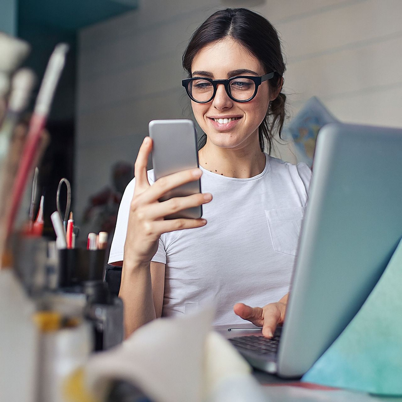 A young woman wearing glasses smiling while looking at her smartphone at a desk with a laptop.