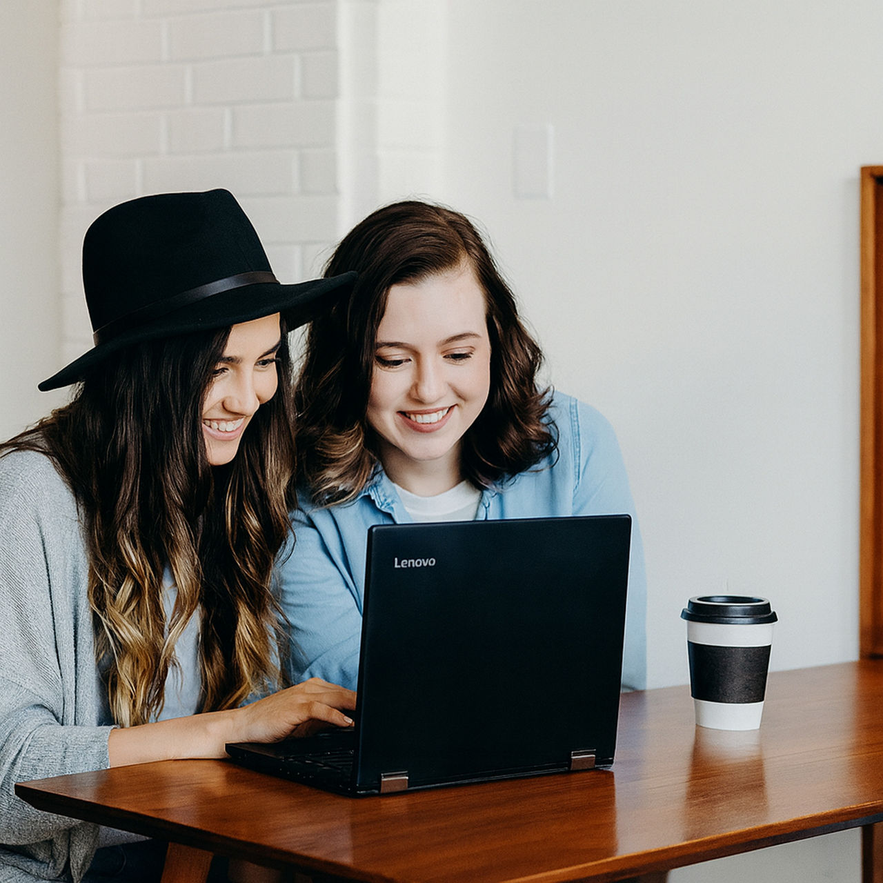 Two young women sitting at a wooden table, smiling while looking at a laptop, with coffee cups nearby.