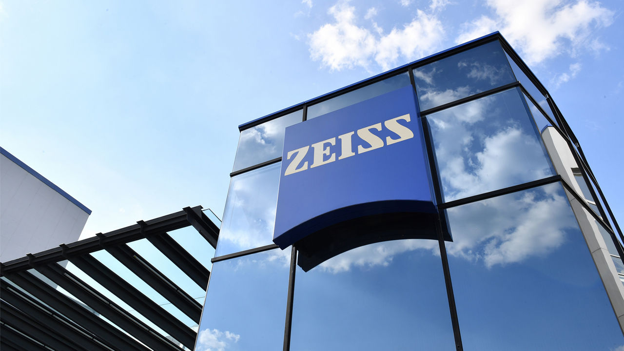 A modern ZEISS office building with a reflective glass facade and a large blue ZEISS logo against a bright, cloudy sky.