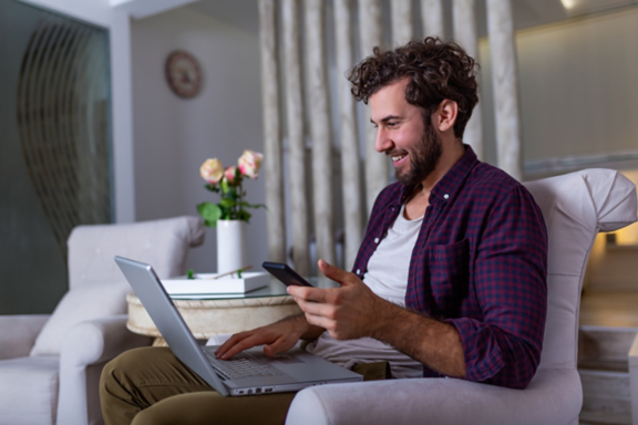 Successful entrepreneur smiling in satisfaction as he checks information on his laptop computer while working in a home office. Young man relaxing on the sofa with a laptop and mobile phone in hands