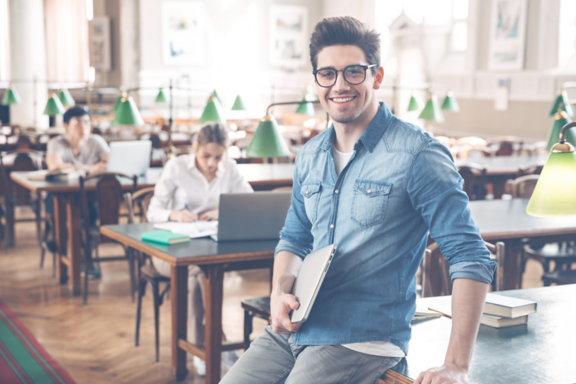 Smiling male student studying at the library, using laptop computer