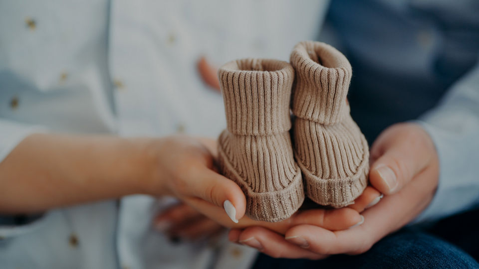 Nahaufnahme von zwei Händen, die ein Paar kleine, beige, gestrickte Babyschuhe umfassen.