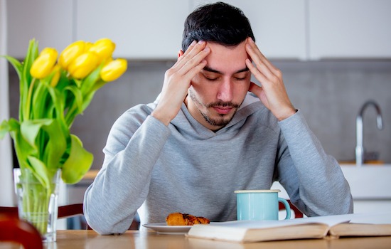 Young tired man sitting at table with cup of coffee or tea and book. Kitchen location.