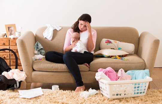 Mother with her baby on her knee. Her head is lowered as she sits on a sofa looking tired and stressed. The room looks untidy with clothes, nappies and other items scattered around the room.