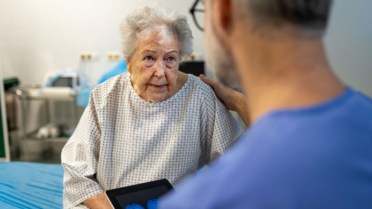 Arzt oder Ärztin spricht mit einer älteren Patientin am Krankenhausbett und zeigt ihr dabei Daten auf einem Tablet