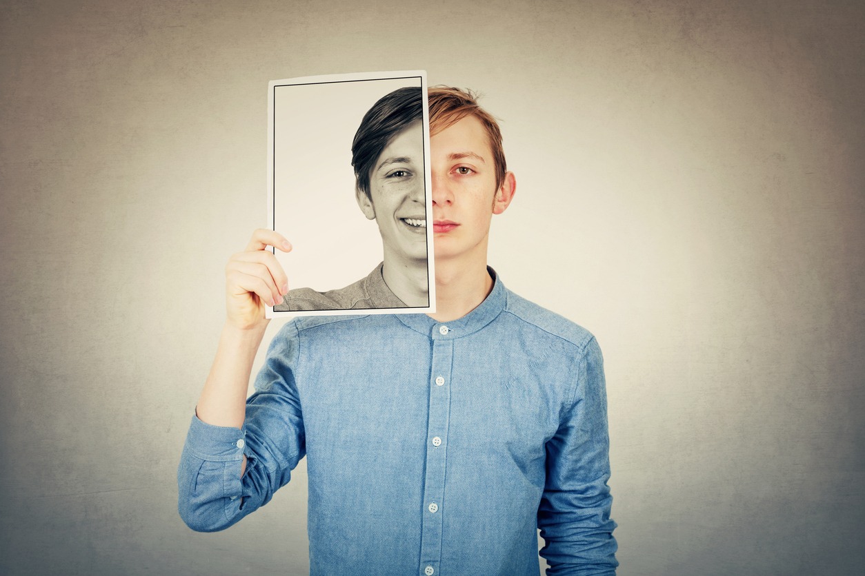 False portrait of a sad boy teenager hiding half face using a photo print paper sheet with a fake happy emotion. Mask for hiding the real expression, create new fake identity, grey wall background.