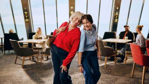 Dwie uśmiechnięte kobiety w salonie Stena Line z widokiem na morze, wygodne miejsca i inni pasażerowie w tle.