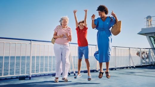Three people are jumping together on the open deck of a ferry or cruise ship. An older woman in a striped shirt, a child in a red shirt, and an adult in a blue dress enjoy a sunny day by the sea. The bright blue sky and ocean create a cheerful vacation atmosphere. A white railing and ship structure frame the scene, emphasizing travel and leisure.
