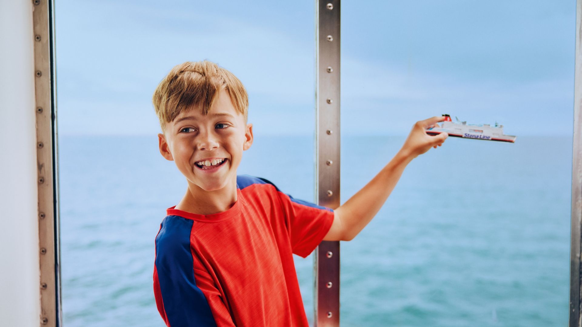A young boy in a red and blue shirt stands by a large window overlooking the ocean, holding up a toy ship. The scene is set on a boat or ferry, with calm blue water visible outside. The boy appears to be enjoying a travel or vacation experience, with a playful and relaxed mood. No visible text or numbers are present in the image.