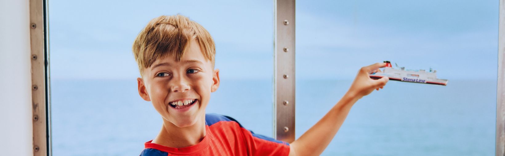 A young boy in a red and blue shirt stands by a large window overlooking the ocean, holding up a toy ship. The scene is set on a boat or ferry, with calm blue water visible outside. The boy appears to be enjoying a travel or vacation experience, with a playful and relaxed mood.