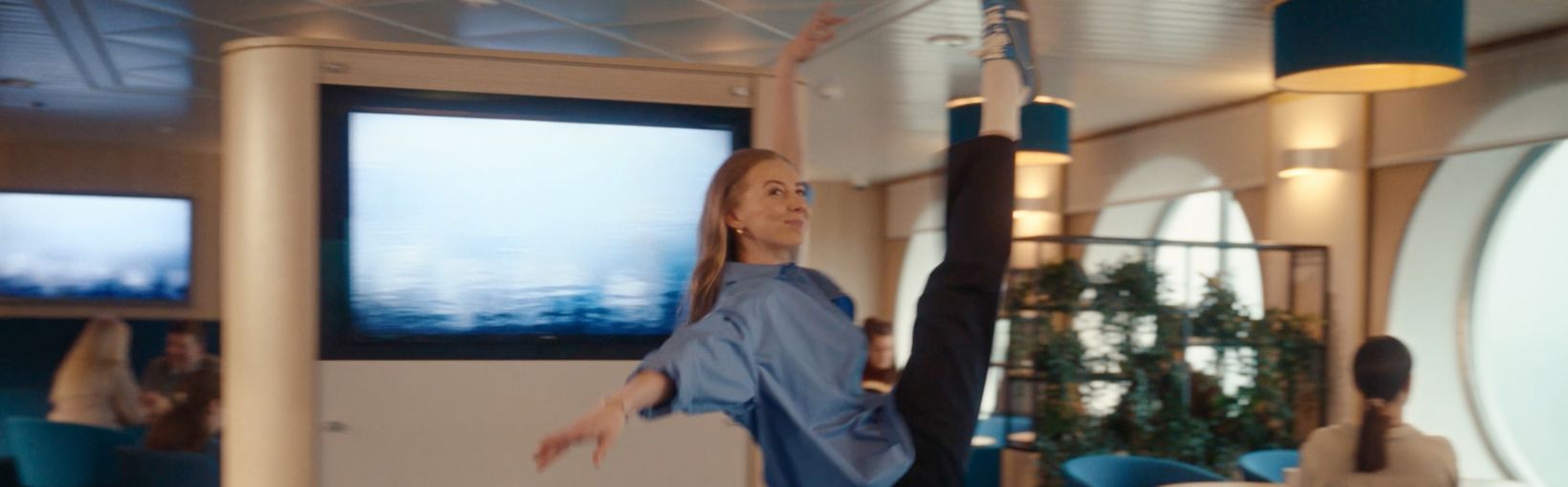 En kvinna utför ett högt danssteg med ena benet sträckt uppåt, i loungemiljö ombord på en färja. Andra passagerare syns i bakgrunden, sittandes vid bord.