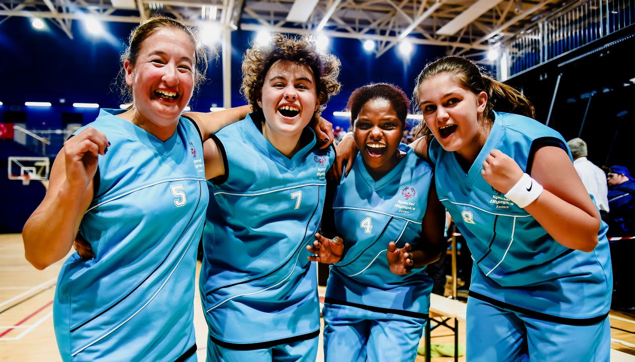 Four women in matching blue basketball uniforms pose together on an indoor basketball court. The group displays camaraderie and excitement, with arms around each other and fists raised in celebration. Visible jersey numbers include 5, 7, and 4. The background features bright stadium lighting and a basketball hoop.