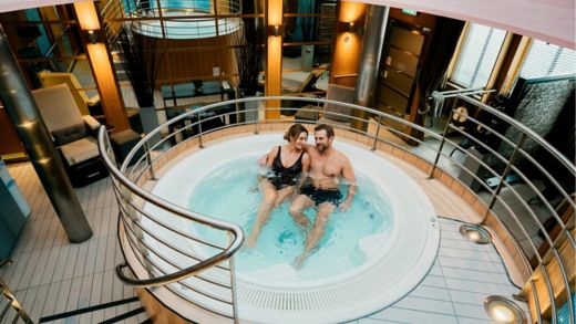 A couple sits together in a circular indoor hot tub, enjoying a relaxing soak. The spa area features warm lighting, metal railings, and modern architectural details. Lounge chairs and large windows with blinds surround the pool, creating an upscale wellness atmosphere. The overall scene conveys comfort, intimacy, and leisure in a luxurious setting.
