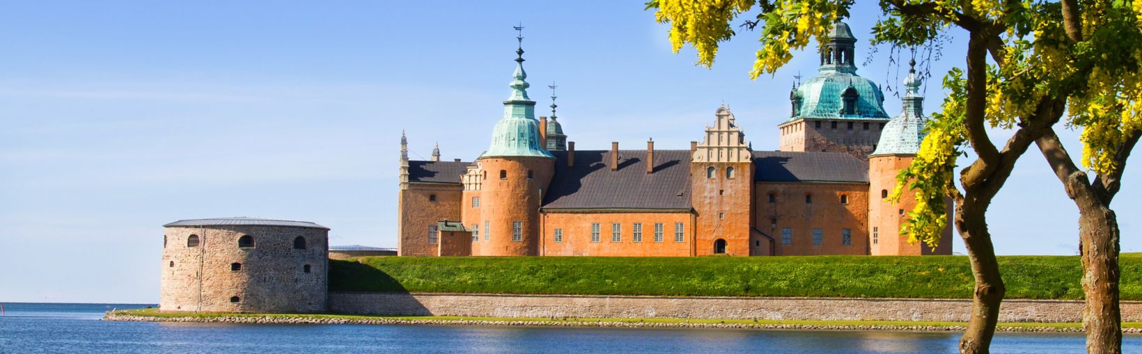 Ūdens ieskauta viduslaiku pils ar sarkanām sienām un ziliem tornīšiem saulainā dienā Kalmarā, Zviedrijā.