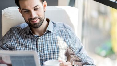 Feeling happy. Positive stylish bearded man is enjoying hot espresso while reading newspaper with smile